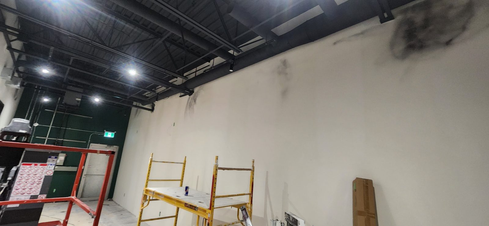 Tall finished drywall wall beneath an open black ceiling with scaffolding in a commercial space
