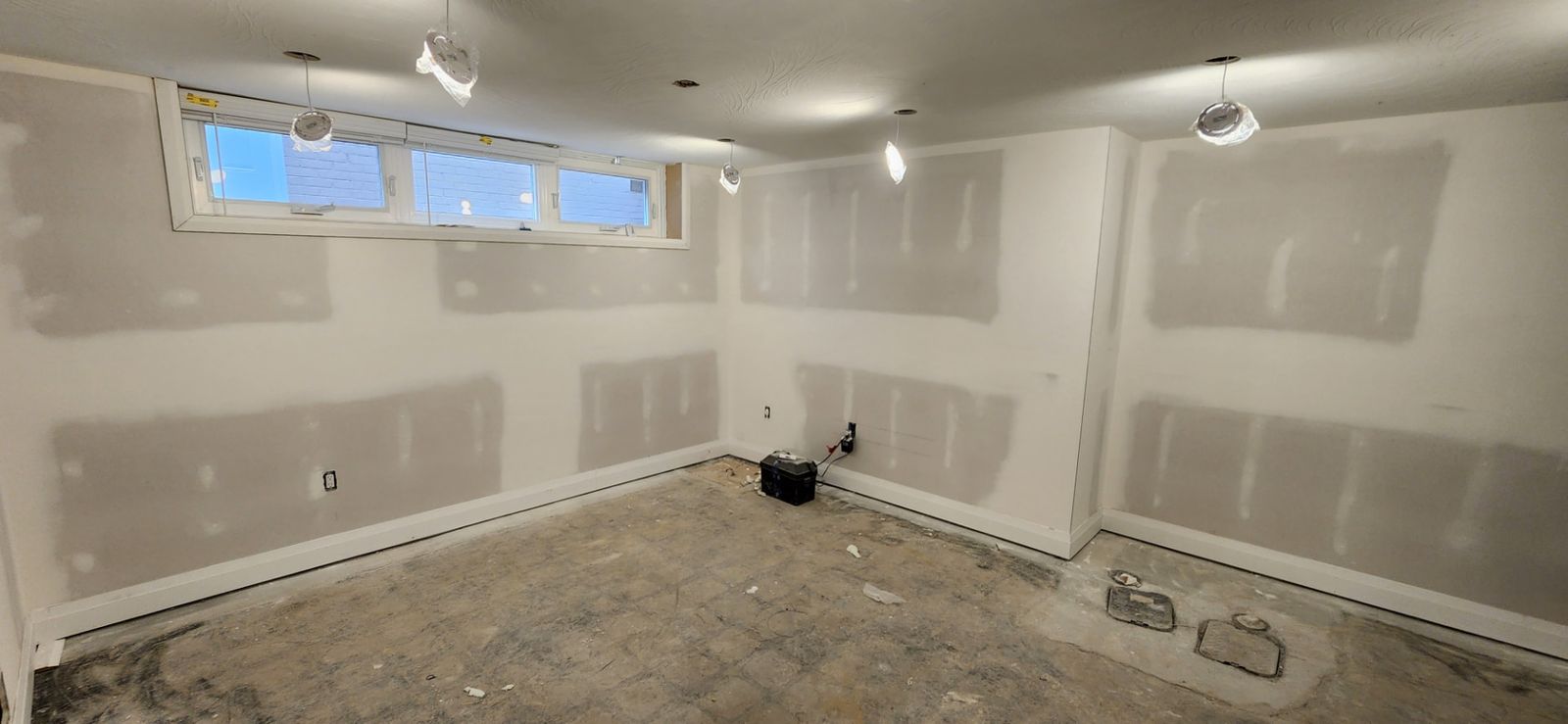 Basement room with patched and sanded drywall walls, new baseboards and work lights near a wide window
