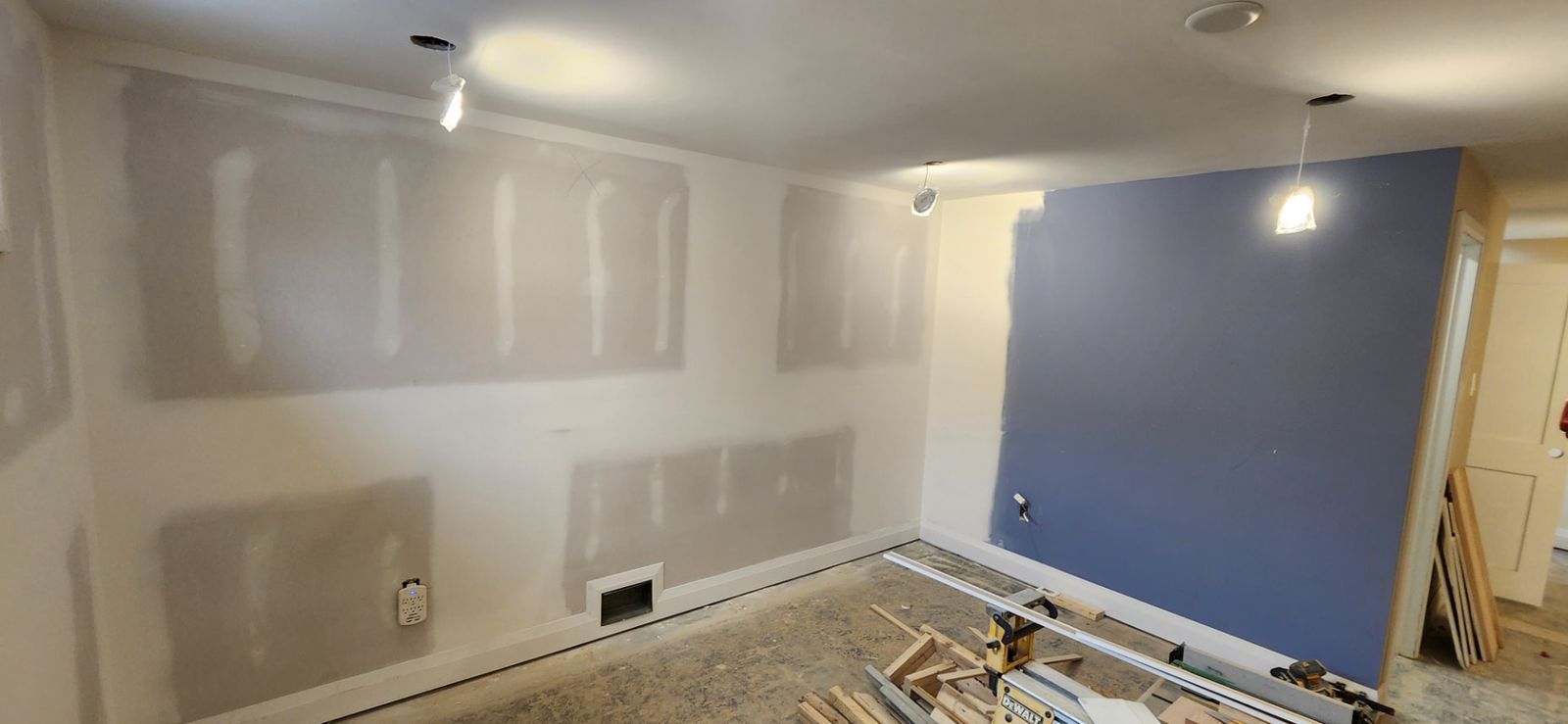 After: Basement recreation room with drywall walls before final coat and a blue painted accent wall under work lights